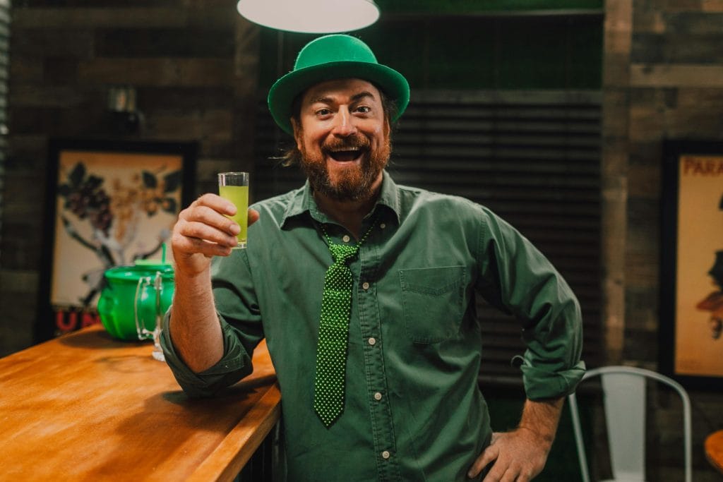 Cheerful man in green attire holding a drink, celebrating St. Patrick's Day indoors.